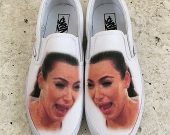 May include: A pair of white canvas slip-on shoes with a printed image of a woman crying. The shoes have the word "Vans" printed on the back.