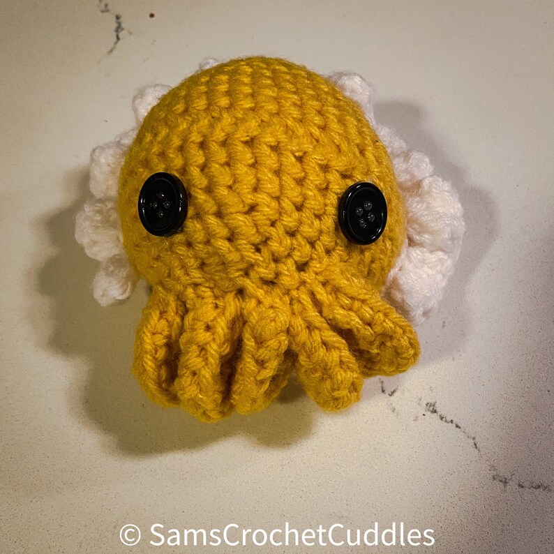K&ouml;nnte beinhalten: Ein geh&auml;keltes Octopus-Amigurumi-Spielzeug mit gelbem K&ouml;rper und wei&szlig;en Tentakeln. Der Octopus hat zwei schwarze Knopfaugen.