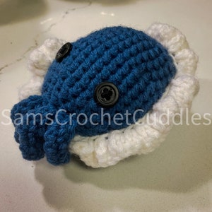 K&ouml;nnte beinhalten: Ein blauer geh&auml;kelter Oktopus mit wei&szlig;en geh&auml;kelten R&uuml;schen um den Kopf. Der Oktopus hat zwei schwarze Kn&ouml;pfe als Augen.