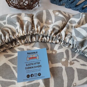 May include: Gray and white geometric patterned fitted cushion cover with elastic edges. The cover is made by Sensible Home Decor and is designed to protect furniture. The text on the tag reads "Sensible Home Decor with Purpose Elastic Fitted Cushion Covers Protect your furniture in style Simple to remove and wash www.etsy.com/shop/SensibleHomeDecor"