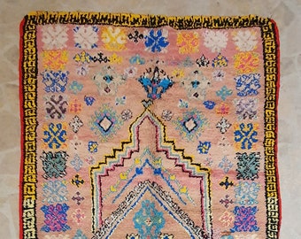 May include: A pink Moroccan rug with a geometric pattern in shades of blue, yellow, green, and red. The rug has a fringe border.