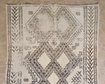 May include: A white rug with a geometric pattern in brown. The pattern is made up of diamonds and squares. The rug has a fringe border.