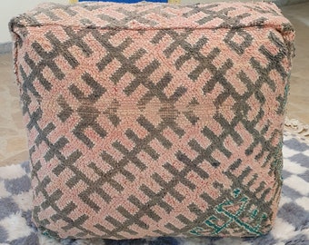 Può includere: Un pouf rosa e grigio con un motivo geometrico. Il pouf è appoggiato su un tappeto a scacchi bianco e grigio.