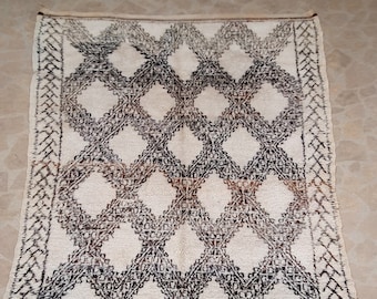 May include: A white Moroccan rug with a geometric pattern in shades of brown and black. The rug has a fringe border.