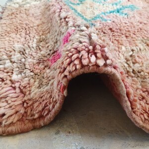 May include: A close-up of a pink and white Moroccan rug with a frayed edge. The rug is folded in half, revealing the underside.