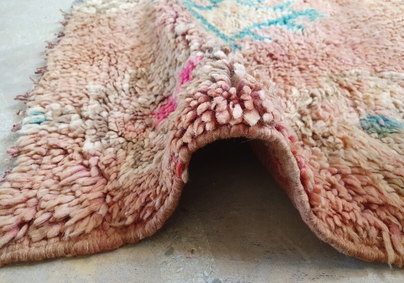 May include: A close-up of a pink and white Moroccan rug with a frayed edge. The rug is folded in half, revealing the underside.