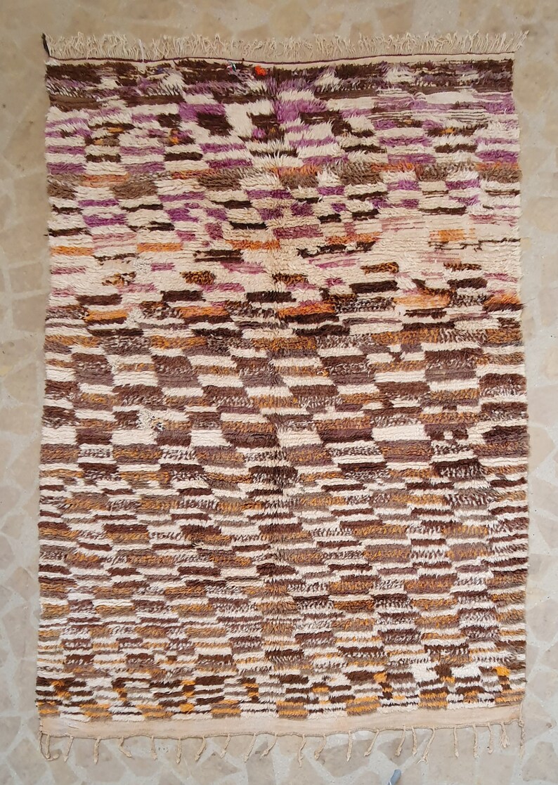 May include: A Moroccan rug with a geometric pattern in shades of brown, white, and pink. The rug has a fringe border.