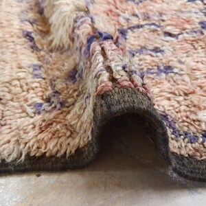 May include: A close-up of a beige, pink, and purple woven rug with a fringe border. The rug is folded in half, showing the underside of the rug.