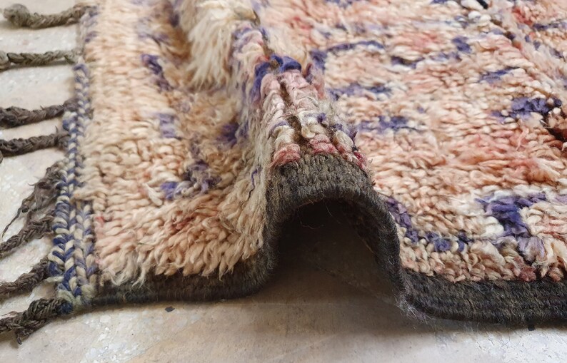 May include: A close-up of a beige, pink, and purple woven rug with a fringe border. The rug is folded in half, showing the underside of the rug.