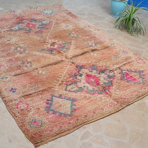 May include: A vintage Moroccan rug with a beige background and intricate patterns in shades of pink, blue, and brown. The rug has a distressed look and is woven with a variety of textures.
