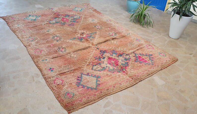 May include: A vintage Moroccan rug with a beige background and intricate patterns in shades of pink, blue, and brown. The rug has a distressed look and is woven with a variety of textures.