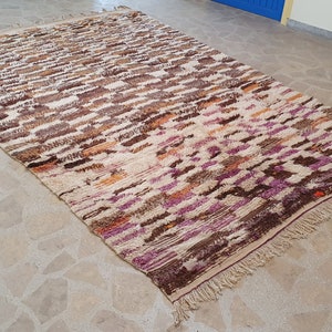 May include: A hand-woven Moroccan rug with a beige background and a colorful geometric pattern in shades of brown, orange, purple, and pink. The rug has a fringe border.