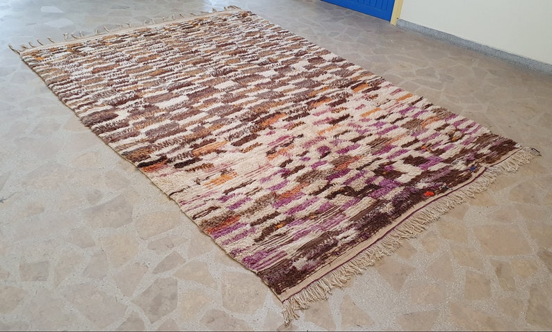 May include: A hand-woven Moroccan rug with a beige background and a colorful geometric pattern in shades of brown, orange, purple, and pink. The rug has a fringe border.