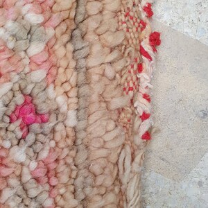 May include: Close-up of a beige and pink woven rug with a red fringe. The rug has a textured, hand-woven look.