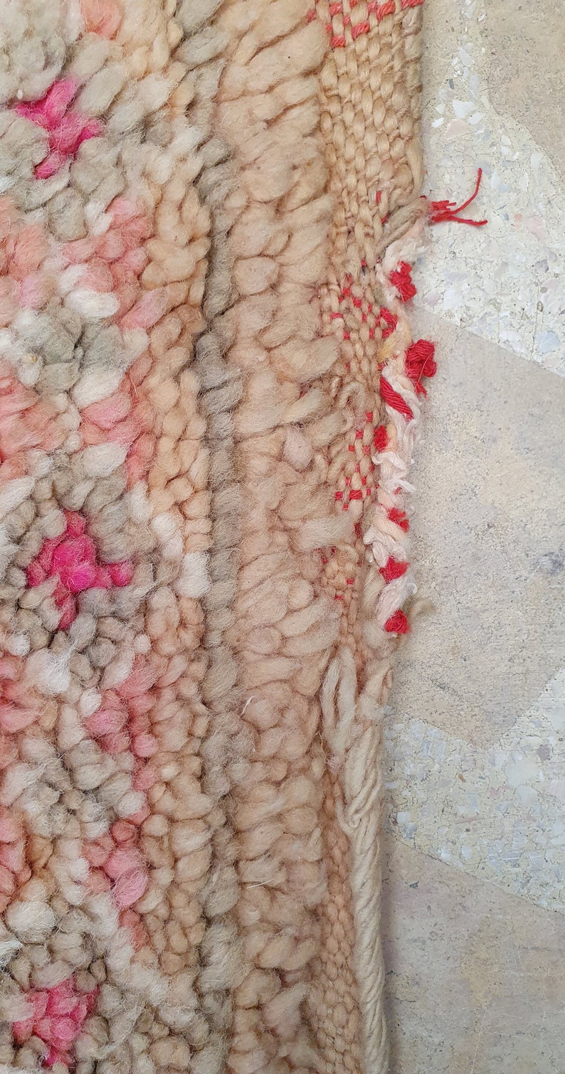 May include: Close-up of a beige and pink woven rug with a red fringe. The rug has a textured, hand-woven look.