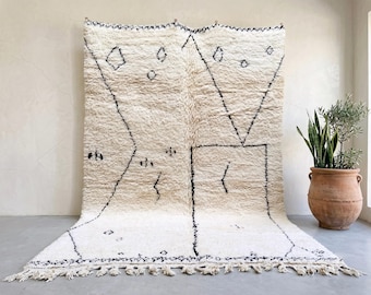 May include: A large, rectangular, off-white area rug featuring a thick, shaggy pile texture. The rug is decorated with black, hand-drawn geometric lines, including triangles, diamonds, and small abstract symbols, finished with a fringed edge.