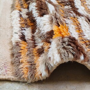 May include: A handwoven rug with a shaggy texture in shades of brown, white, and orange. The rug has a fringe border and is made of natural fibers.