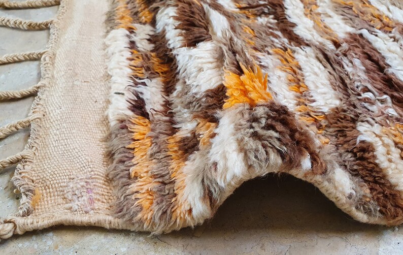 May include: A handwoven rug with a shaggy texture in shades of brown, white, and orange. The rug has a fringe border and is made of natural fibers.
