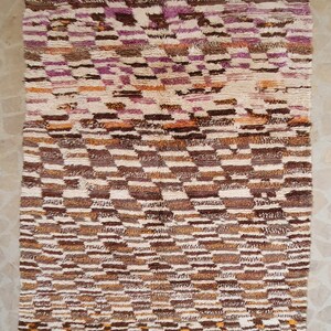 May include: A Moroccan rug with a geometric pattern in shades of brown, white, and pink. The rug has a fringe border.