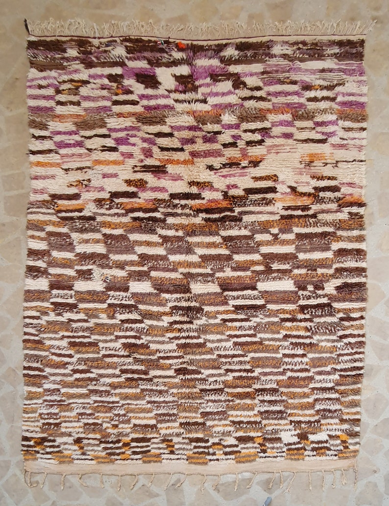 May include: A Moroccan rug with a geometric pattern in shades of brown, white, and pink. The rug has a fringe border.