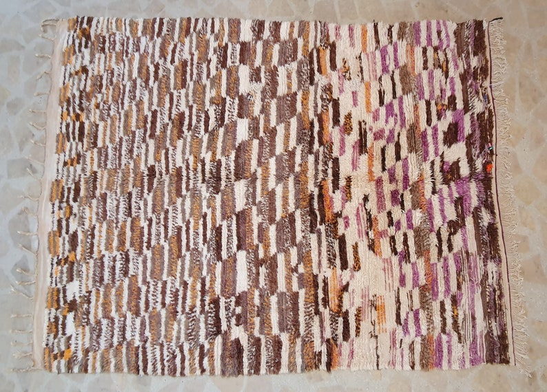 May include: A rectangular rug with a geometric pattern in shades of brown, white, and purple. The rug has a fringe border.
