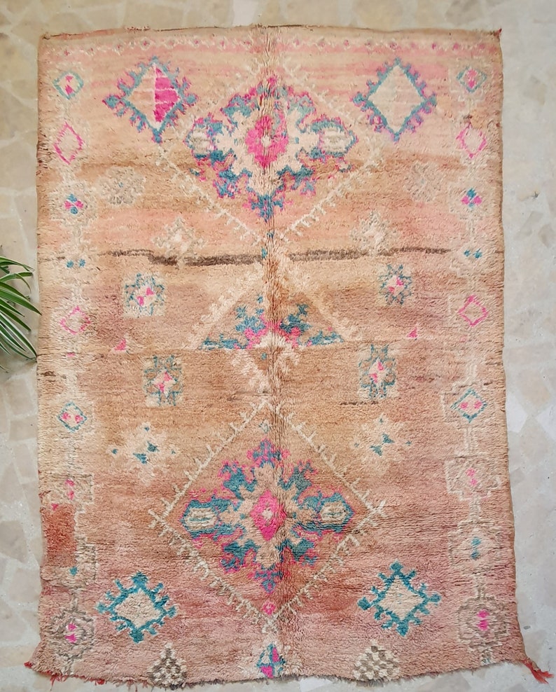 May include: A vintage Moroccan rug with a beige background and intricate patterns in shades of pink, blue, and white. The rug features a central diamond shape with smaller diamond patterns throughout.