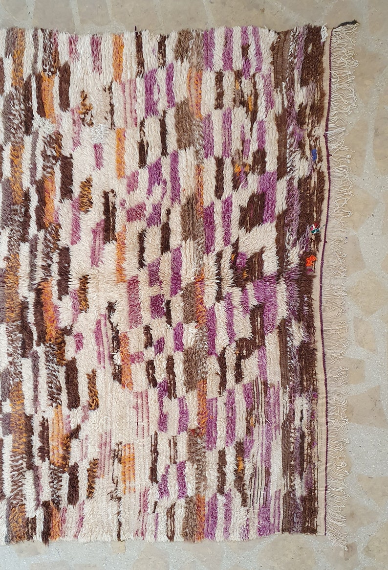 May include: A white, brown, orange, and purple Moroccan rug with a geometric pattern. The rug has a fringe border.