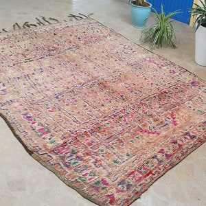 May include: A vintage Moroccan rug with a beige background and intricate patterns in shades of pink, purple, and green. The rug has a fringed border and is woven with a variety of textures.