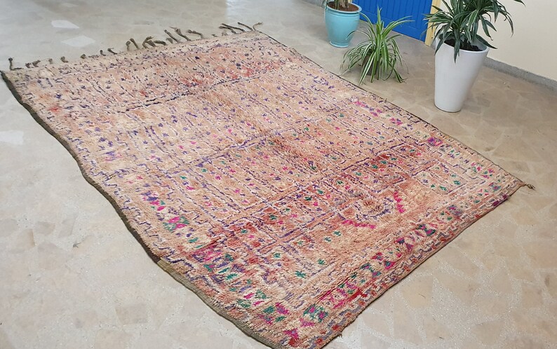 May include: A vintage Moroccan rug with a beige background and intricate patterns in shades of pink, purple, and green. The rug has a fringed border and is woven with a variety of textures.