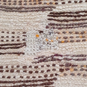 May include: Close-up of a woven rug with a white background and brown, orange, and purple accents. The rug has a geometric pattern with stripes and dots.