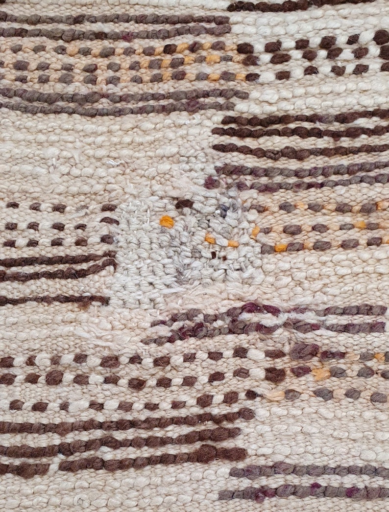 May include: Close-up of a woven rug with a white background and brown, orange, and purple accents. The rug has a geometric pattern with stripes and dots.