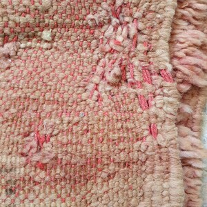 May include: Close-up of a woven rug with a pink and beige pattern. The rug has a frayed edge and is made of a soft, textured material.