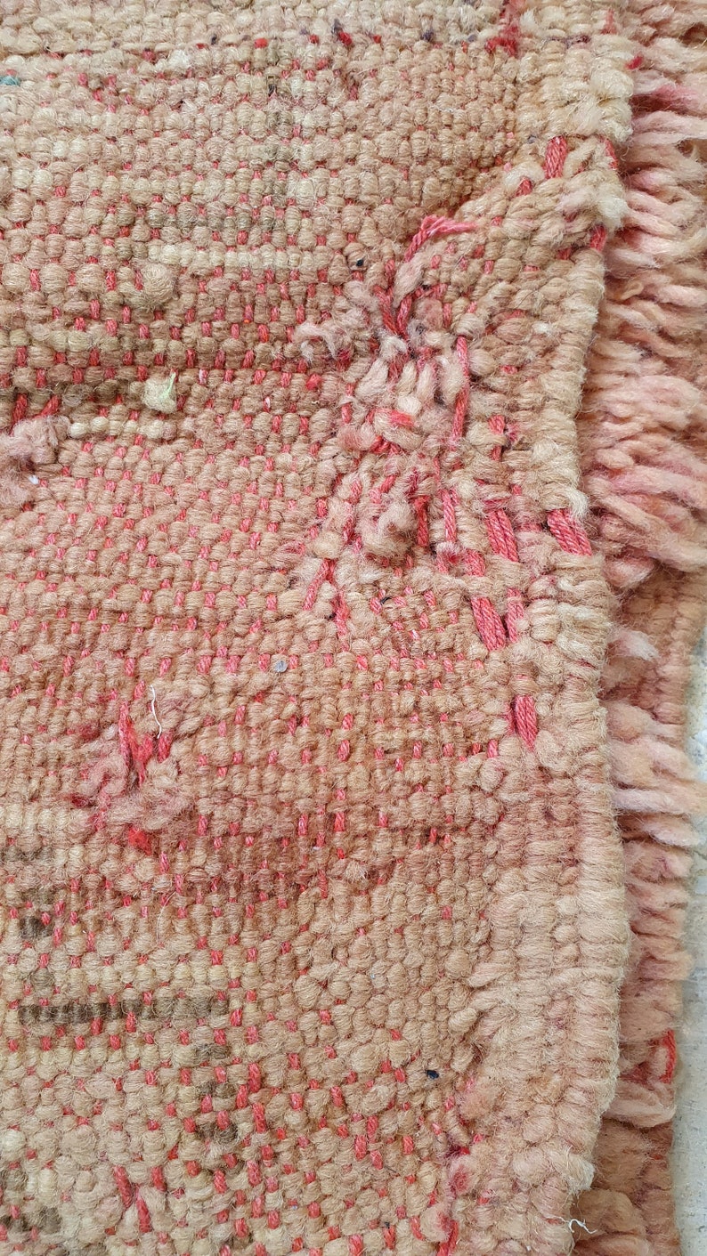May include: Close-up of a woven rug with a pink and beige pattern. The rug has a frayed edge and is made of a soft, textured material.