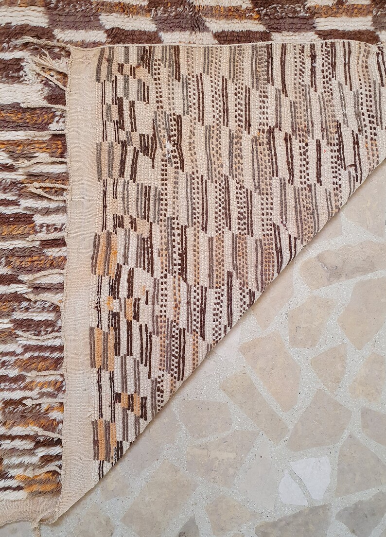 May include: Close-up of the back of a brown and white woven rug with a geometric pattern. The rug is folded in half, revealing the white backing.