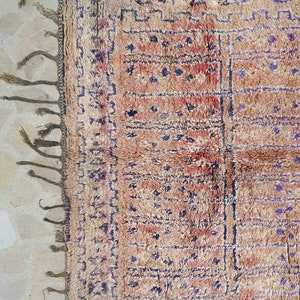 May include: A vintage Moroccan rug with a beige background and a geometric pattern in shades of blue and red. The rug has fringe on all sides.
