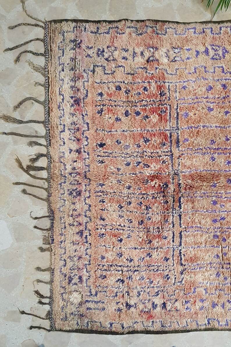 May include: A vintage Moroccan rug with a beige background and a geometric pattern in shades of blue and red. The rug has fringe on all sides.