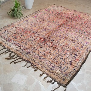 May include: A vintage Moroccan rug with a beige background and intricate patterns in shades of purple, red, and green. The rug has a fringe border and is made of wool.
