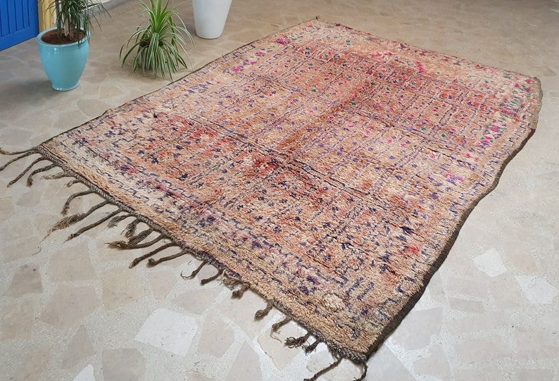May include: A vintage Moroccan rug with a beige background and intricate patterns in shades of purple, red, and green. The rug has a fringe border and is made of wool.