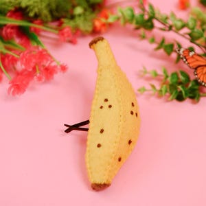 May include: A yellow felt banana hair clip featuring brown stitched details at the top and bottom ends, with small brown embroidered dots scattered across the surface. The banana is attached to a black metal hair clip visible on the side.