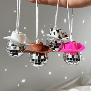 May include: Four spherical disco ball ornaments, each topped with a miniature cowboy hat. The hats are white, brown, black and white cow print, and bright pink. Each ornament hangs from a white string, featuring reflective mirrored glass tiles.