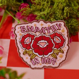 May include: A light pink embroidered patch featuring three red roses with green leaves and the text "EMPATHY IS HOT" in pink lettering. The patch has a scalloped brown border and is decorated with small gold star accents throughout the design.