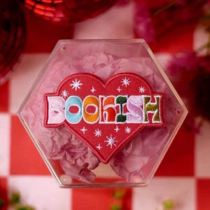 May include: A heart-shaped embroidered patch with the word BOOKISH in multicolored letters, accented by white stitched stars. The patch is contained within a clear hexagonal plastic box filled with light pink tissue paper, displaying the design.
