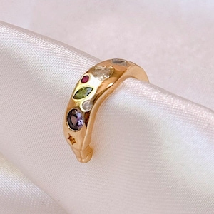 Op de afbeelding: Een goudkleurige oormanchet met een reeks kleine, veelkleurige edelstenen ingelegd langs het buitenoppervlak. De stenen omvatten een ronde paarse edelsteen, een groene edelsteen in marquise-slijpvorm, een kleine rode edelsteen en diverse heldere witte accenten.