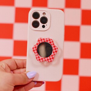 May include: A light pink smartphone case featuring a circular, flower-shaped grip attachment on the back. The grip has a red and pink checkered pattern and a central circular mirror. The case includes a cutout for a multi-lens camera system.