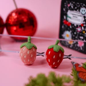 Puede incluir: Dos horquillas de fieltro con forma de fresa. Una fresa es de color rosa claro con semillas bordadas en blanco y un tallo verde, mientras que la otra es de color rojo brillante con semillas bordadas en blanco y un tallo verde. Clips negros.