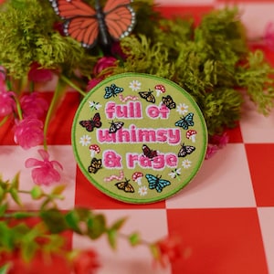 May include: A circular embroidered patch with a light green background featuring the pink text full of whimsy & rage. The design includes small colorful butterflies, white flowers, red and white mushrooms, and pink worms scattered across the surface.