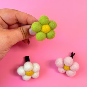 May include: Three felt flower hair clips in the image. One is held by a hand, featuring a green flower with a yellow center. The other two are white and pink flowers with yellow centers, all on a pink background.