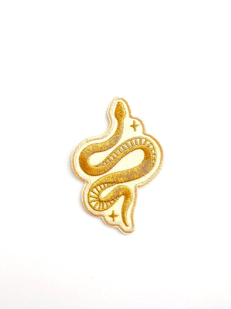 Op de afbeelding: Een geborduurde patch met een kronkelende slang in metallic gouddraad op een cr&egrave;mekleurige achtergrond. Het ontwerp bevat kleine vierpuntige steraccenten boven en onder de slang, met een gestikte rand die de vorm van de slang volgt.