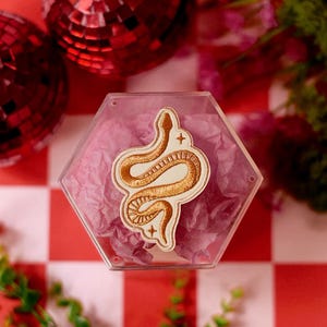 May include: A hexagonal clear plastic container filled with light purple tissue paper, featuring an embroidered patch on the lid. The patch displays a gold-colored snake in a coiled position, accented by two small four-pointed star symbols.
