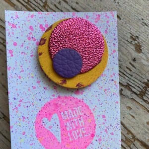 May include: A round, yellow leather pin with a pink and purple leather circle on top. The pin is on a white card with pink and purple paint splatters. The card also has a pink circle with a white heart and the text "Made with Love".
